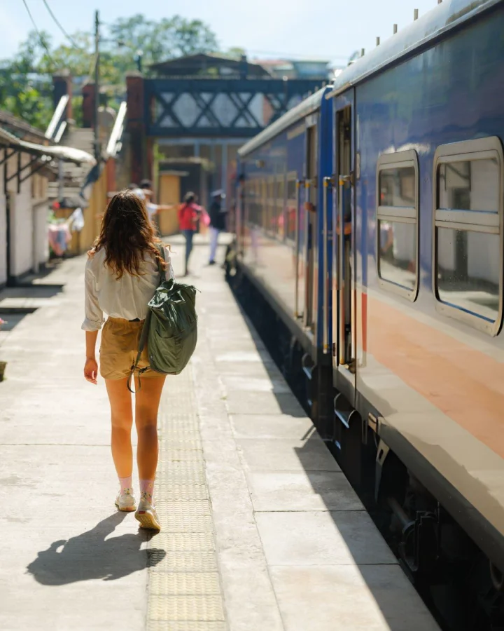 Woman with a backpack walking beside a blue train at Ella railway station in Sri Lanka