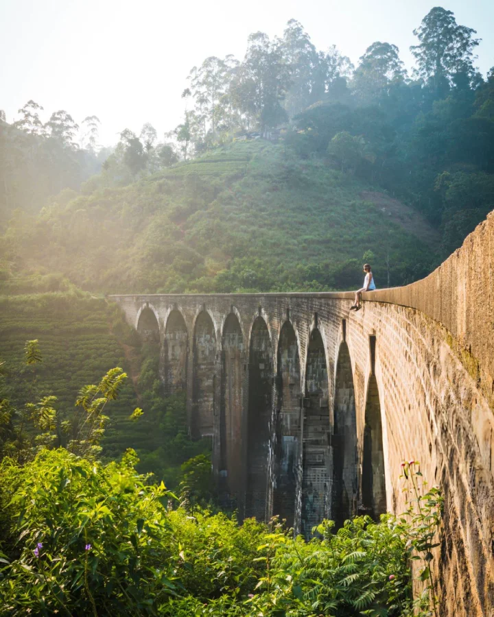 Young woman sitting on Nine Arch Bridge at sunrise, surrounded by green hills